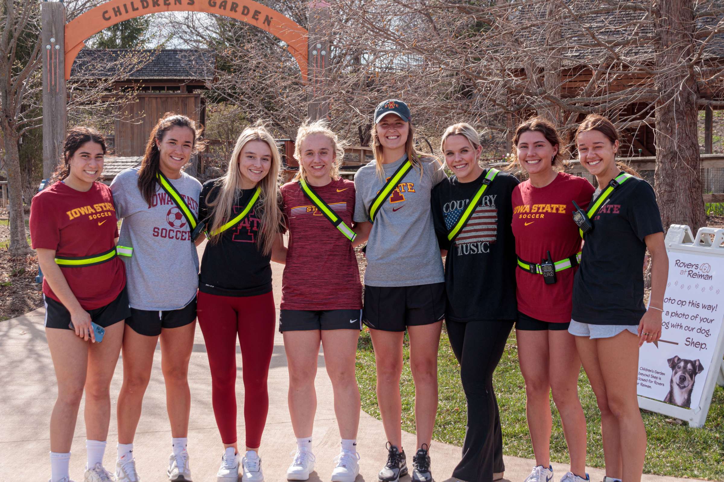 Eight Iowa State women's soccer players stand before the Children's Garden, smiling. These volunteers are excited to volunteer at Rovers at Reiman.