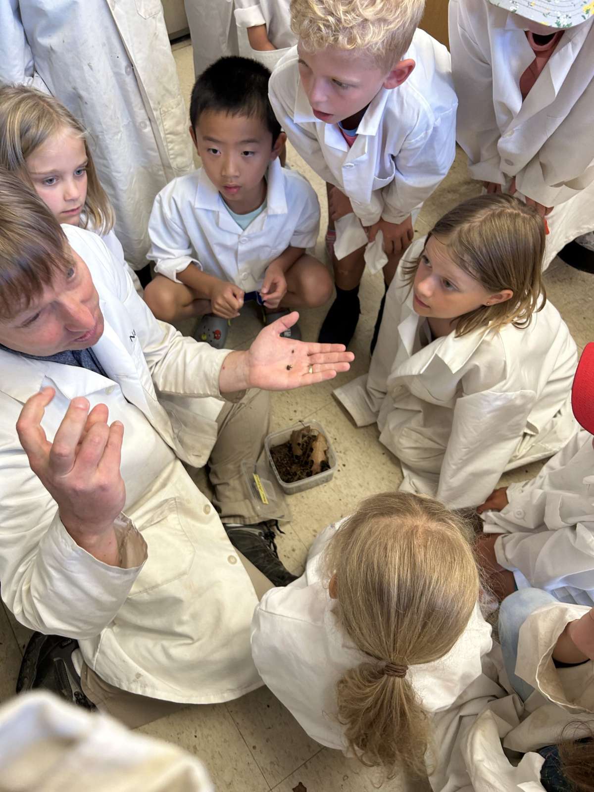 children in lab coats gather around an entomologist in the lab